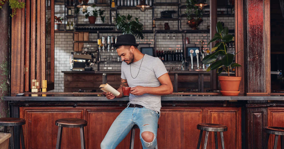 portrait of trendy man sitting at a cafe counter reading a book and drinking coffee